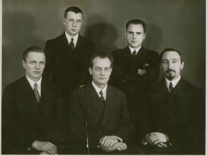 Fotoateljė „Zinaida“. Profesoriai V. Mykolaitis-Putinas, S. Šalkauskis ir kiti. Vincas Mykolaitis-Putinas (sėdi centre), Stasys Šalkauskis (sėdi dešinėje), tekstologas, vertėjas Aleksandras Žirgulys (stovi kairėje), kiti du nežinomi asmenys. Apie 1937–1939 m. Maironio lietuvių literatūros muziejus. Naudojimo teisės: PD. Nuotrauka iš Europeanos