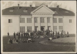 Kauno meno mokyklos dėstytojai ir mokiniai prie mokyklos pastato. Ant suolelio (iš kairės) sėdi: 5) Antanas Žmuidzinavičius, 6) Kajatonas Sklėrius, 7) Domicelė Tarabildaitė-Tarabildienė, (?) 8) Jonas Mackevičius. XX a. 3 deš. ČDM M 1-9-6/27. Nacionalinis M. K. Čiurlionio dailės muziejus. Naudojimo teisės: CC BY-NC. Nuotrauka iš LIMIS