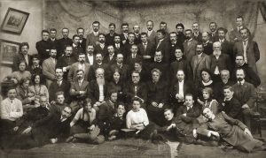 Aleksandras Jurašaitis. Lietuvių mokslo draugijos suvažiavimas 1912 m. Iš kairės: antroje eilėje pirmas – Stasys Šilingas, antra – Emilija Šilingienė, trečias – Juozas Vokietaitis, penktas – Gabrielius Landsbergis-Žemkalnis, šešta – Sofija Kymantaitė-Čiurlionienė, septintas – Jonas Basanavičius, aštuntas – Kazimieras Prapuolenis, devintas – Antanas Vileišis, dešimta – Emilija Vileišienė, dvyliktas – Donatas Malinauskas; trečioje eilėje penktas – Martynas Yčas, aštunta – Liudvika Didžiulienė, dešimtas – Fabijonas Kemėšis. Iš dešinės: ketvirtoje eilėje pirmas – Mykolas Sleževičius, šeštas – Kazys Grinius; penktoje eilėje trečias – Jonas Vileišis, ketvirtas – Antanas Žmuidzinavičius. Lietuvos nacionalinis muziejus. Naudojimo teisės: CC BY-SA 2.5. Nuotrauka iš Vikipedijos